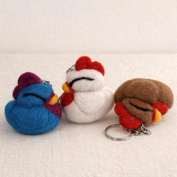 Three needle felted sleeping chicken keychains in blue, white, and brown with red details and metal rings