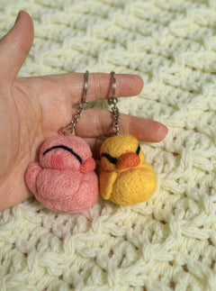 Two needle felted bird keychains in pink and yellow wool held in palm, showing size scale