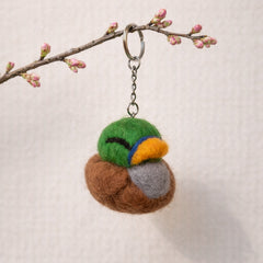 Needle felted mallard bird keychain in green, brown, gray, and gold wool hanging from a branch with pink buds
