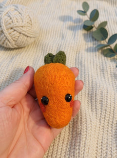 Needle felted orange carrot with green leaves and black eyes held in palm of hand