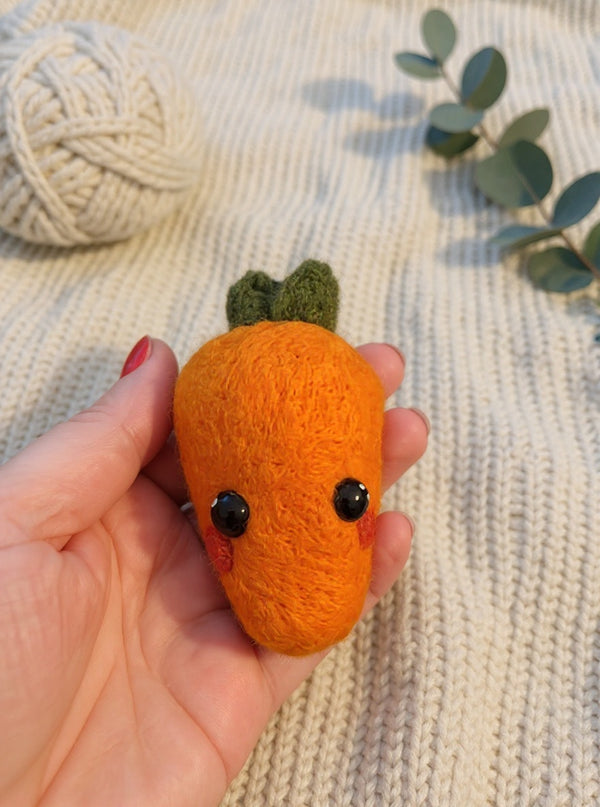 Needle felted orange carrot with green leaves and black eyes held in palm of hand