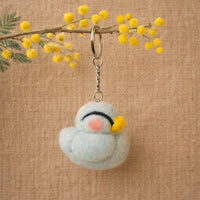Needle felted light blue chicken keychain with white wool body, pink cheeks, yellow flower details, hanging from a branch.