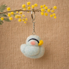 Needle felted light blue chicken keychain with white wool body, pink cheeks, yellow flower details, hanging from a branch.